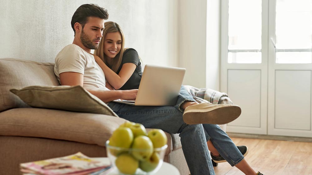 Young couple searches for something in internet, man types in the inquiry while blond woman leans on his shoulder and smiles, unfocused bowl of green apples on the front view