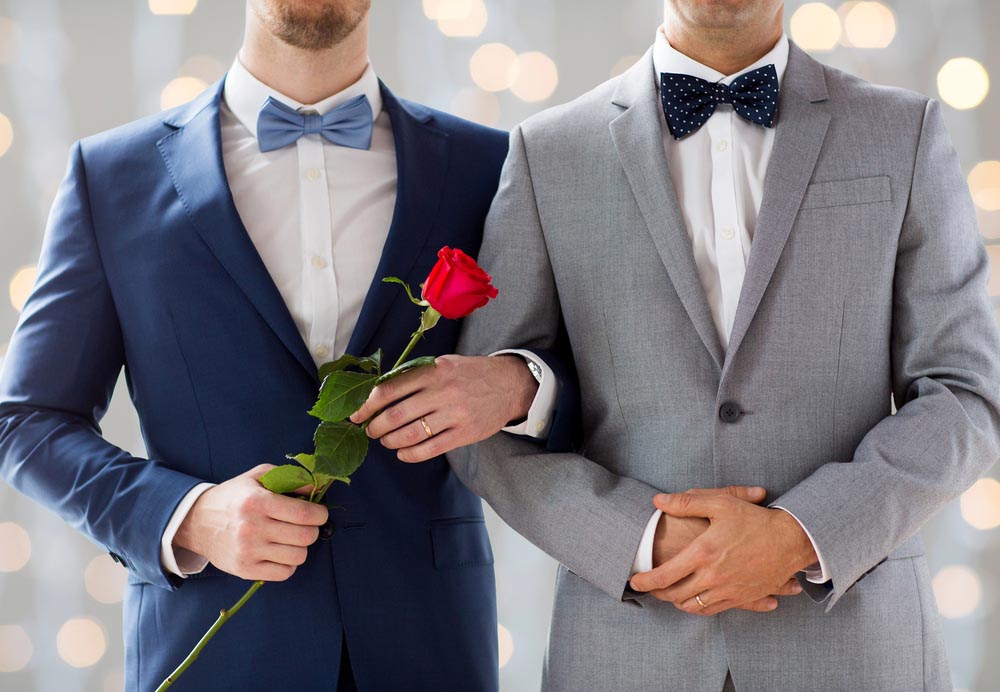 people, homosexuality, same-sex marriage and love concept - close up of happy male gay couple with red rose flower holding hands on wedding over holidays lights background