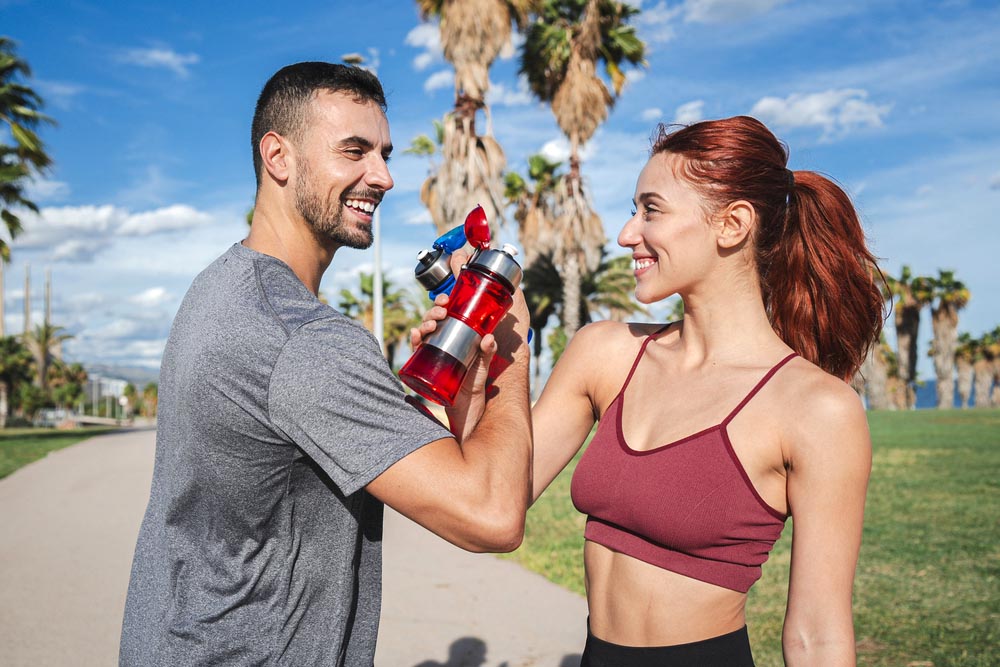 Sport couple toasting and drinking water while resting after run or workout. Trainer man and strong woman, smiling and clinking supplement bottles of having a break and refreshing during training. High quality photo